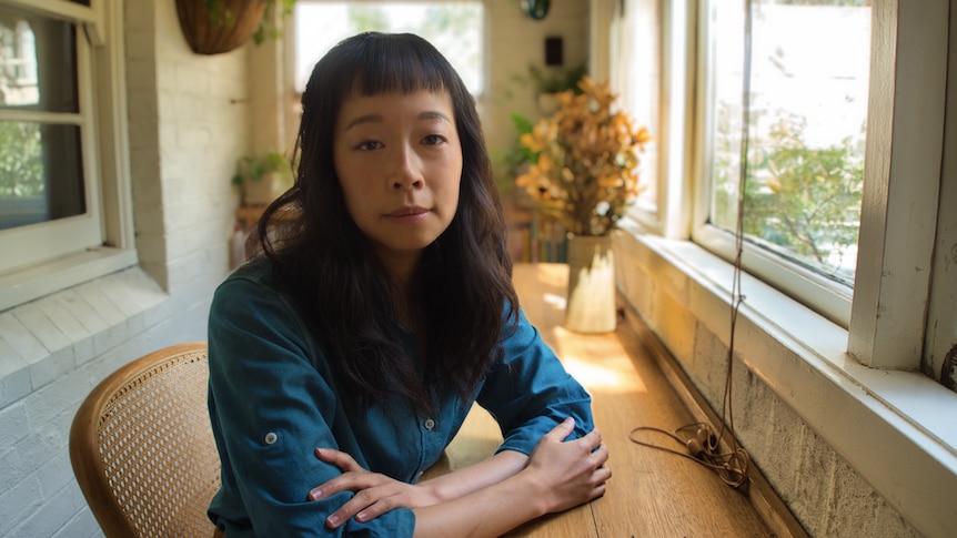 An Asian Australian woman sits at a desk in sunlight, her arms folded in front of her