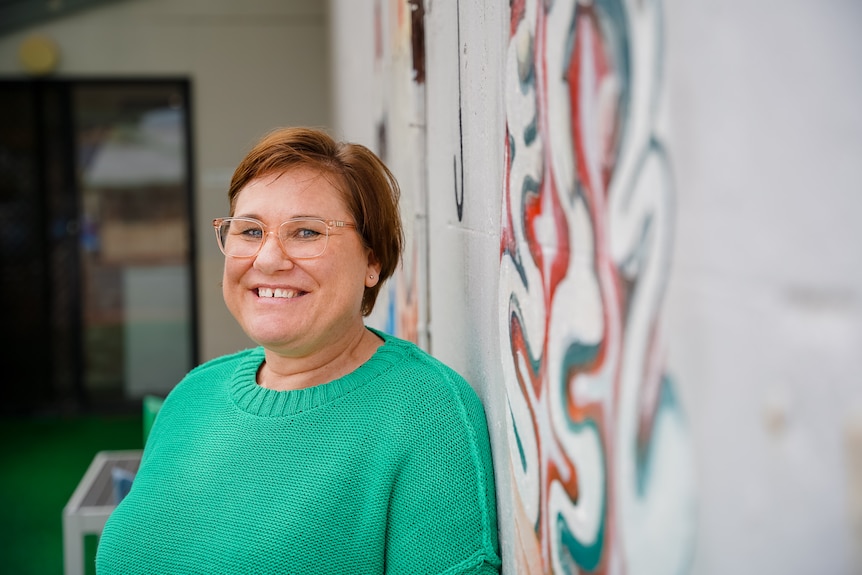 A woman wearing a green jumper and glasses leans against a painted mural of an ice cream cone, smiling.