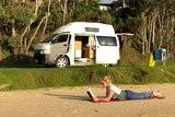 A traveller reads on the beach with her campervan on the foreshore surrounded by trees.