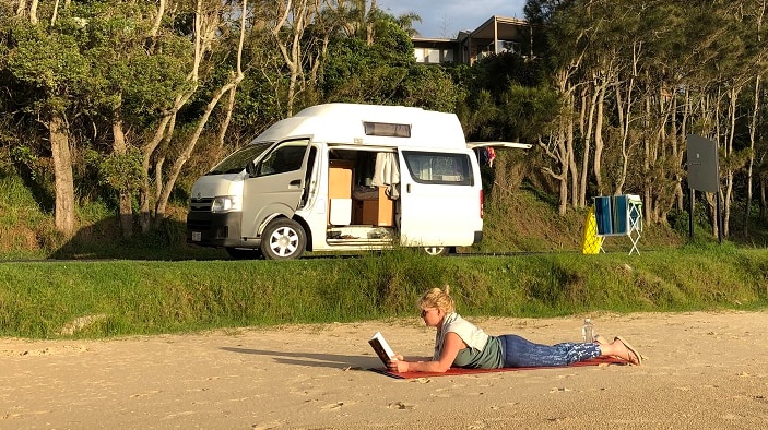 A traveller reads on the beach with her campervan on the foreshore surrounded by trees.