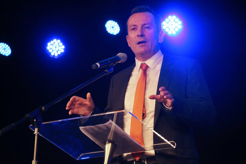 A mid shot of Mark McGowan talking into a microphone at a pedestal with lights behind him.