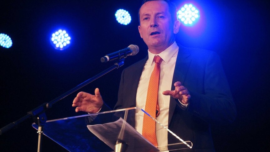 A mid shot of Mark McGowan talking into a microphone at a pedestal with lights behind him.