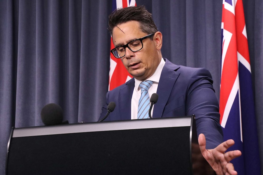 A mid shot of WA Treasurer Ben Wyatt standing in front of two flags at a media conference looking down with an arm outstretched.