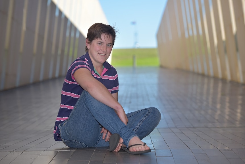 A woman in jeans and shirt sitting and smiling at the camera with a blurred corridor in the background
