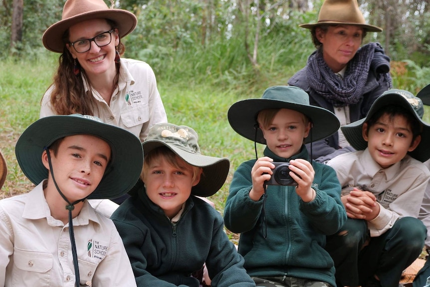 Four students wear a green hat and khaki uniform, seated on a mat in the bush with the principal and another teacher behind them