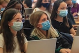 Students sit side by side in a lecture hall, all wearing surgical masks.
