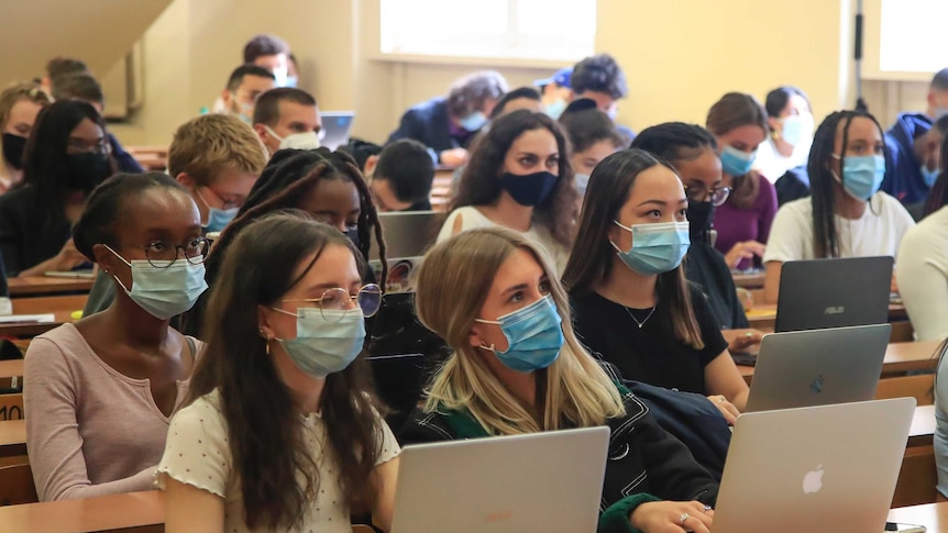 Students sit side by side in a lecture hall, all wearing surgical masks.