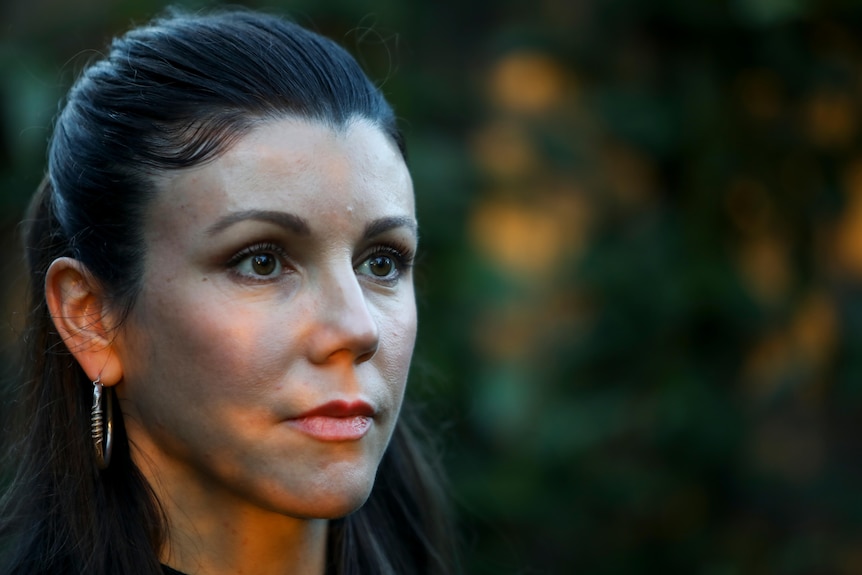 Dappled light falls on the face of a woman with dark hair pulled back in a half-up half-down style staring into the distance