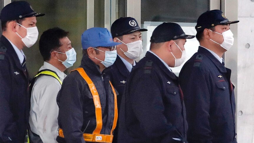 Carlos Ghosn wearing workers outfit and a mask walks among detention centre staff.