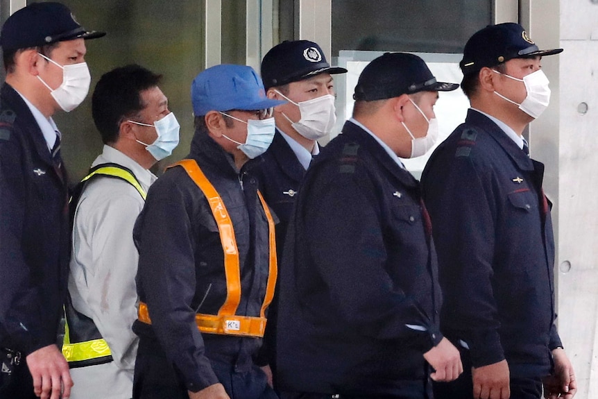 Carlos Ghosn wearing workers outfit and a mask walks among detention centre staff.