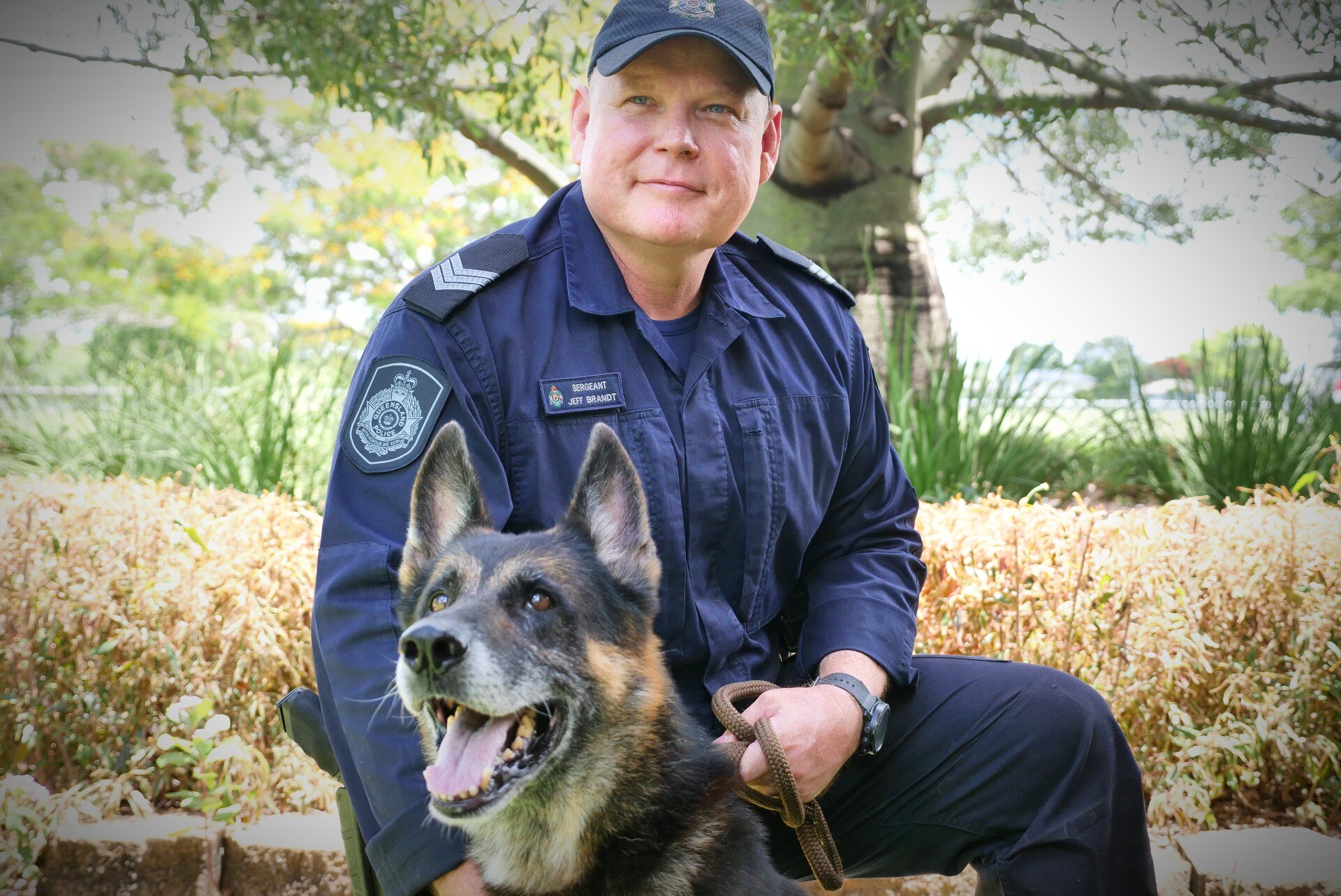 A police officer kneeling down in front of a garden with a German Shepard 