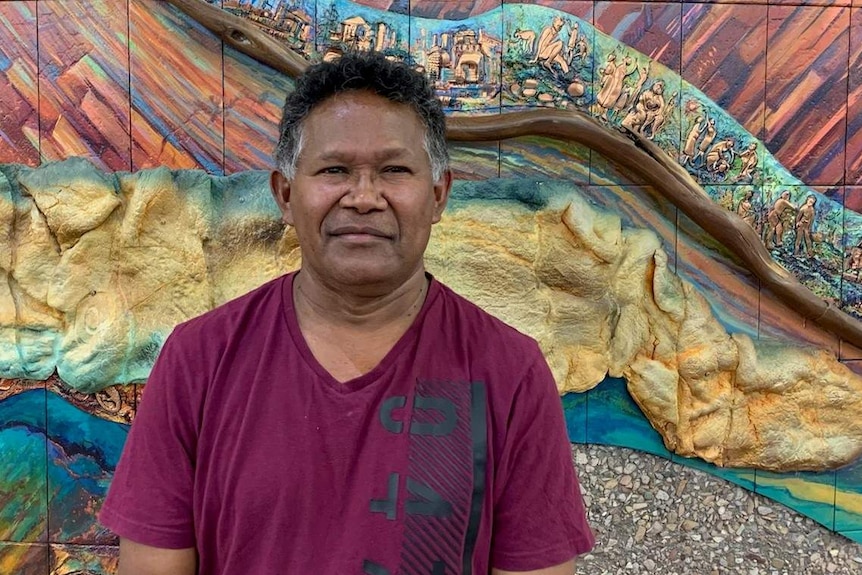 An Indigenous man stands before a mural.