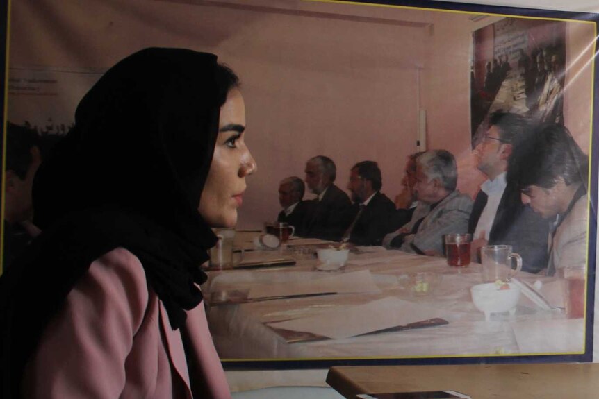 A woman wearing a headscarf speaks in front of a poster of men.