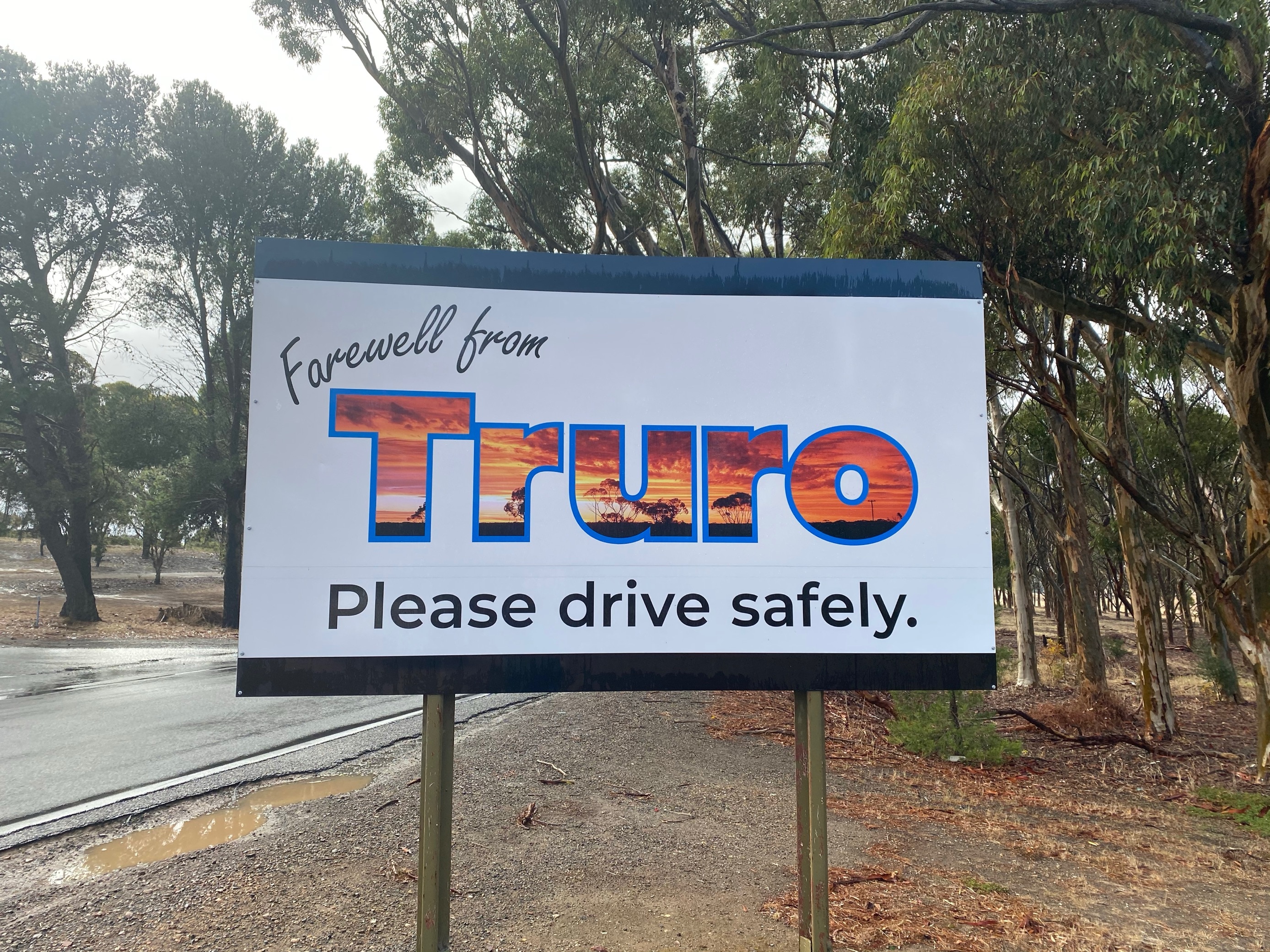 A white sign saying farewell from Truro in front of a wet road.