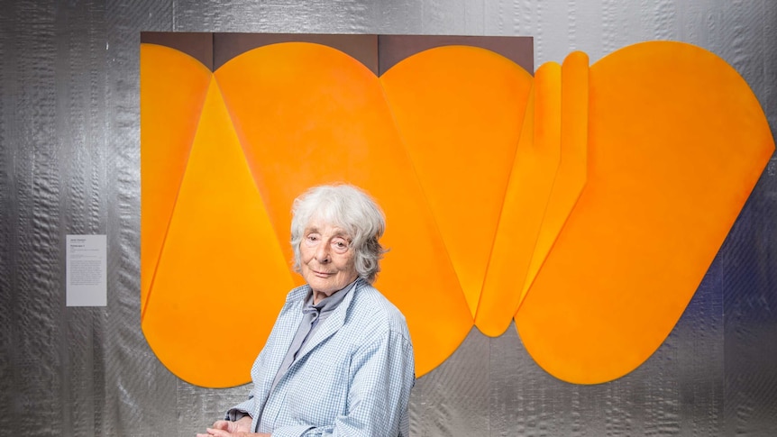 An older woman stands in front of an abstract painting on an oddly shaped canvas
