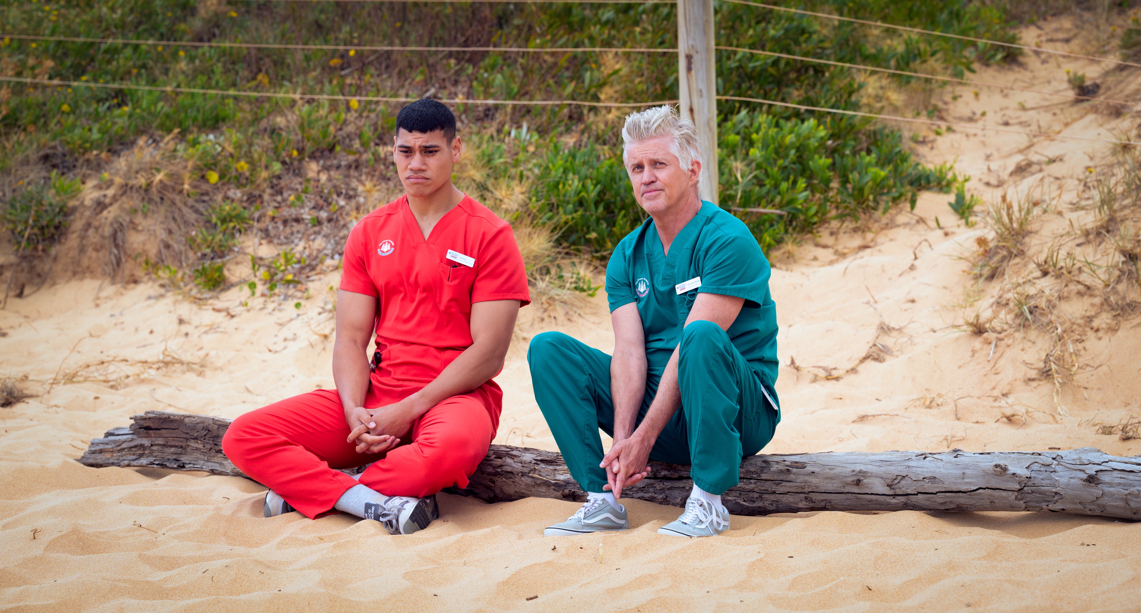 A TV still of Ioane Sa'ula, mid-20s, Samoan Australian, and Dan Wyllie, 56, sit together on a beach, looking sombre.