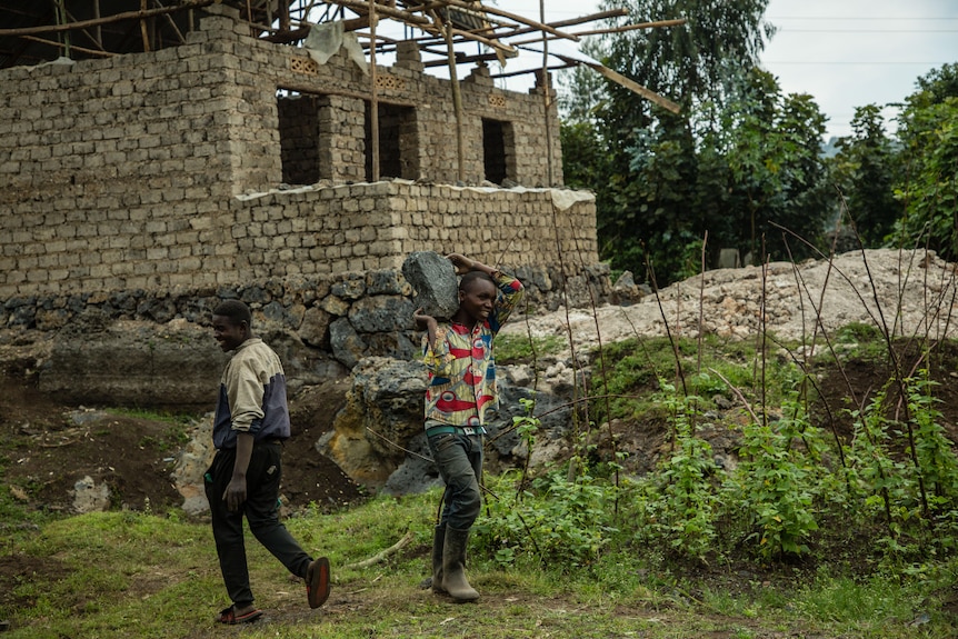 Emmanuel carries a large boulder on his shoulders, he wears a bright shirt.