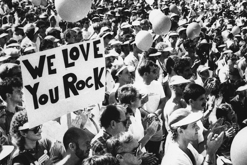 Black and white image og big crowd, Rock Hudson's fans, holding a board 'We love you Rock'.