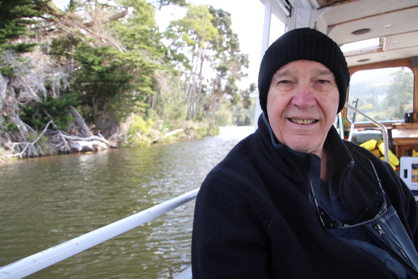 Dean Hohn, wearing a black jumper and black beanie sits on a boat on a river with trees visible on the bank