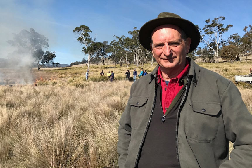 Professor David Bowman stands in grasslands with smoke behind him