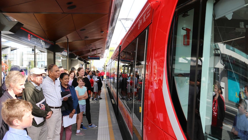 Crowd of people standing on a light rail platform waiting to get on the red tram in front of them.