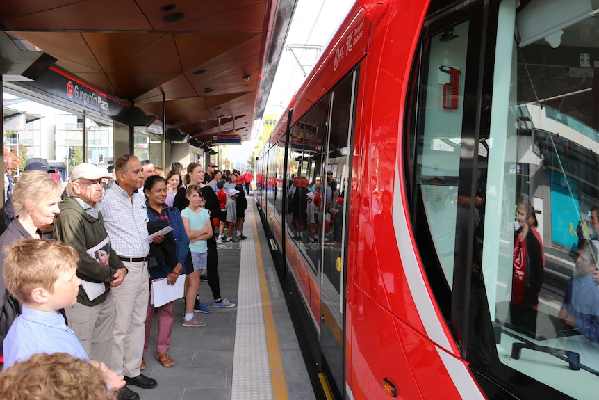 Crowd of people standing on a light rail platform waiting to get on the red tram in front of them.