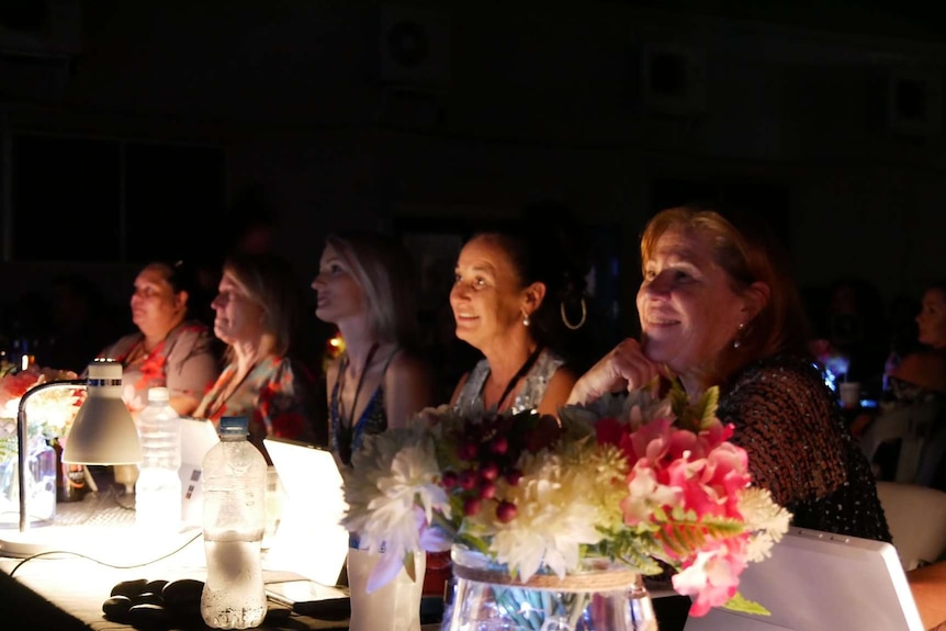 Several female judges sit at a long table looking at a stage.