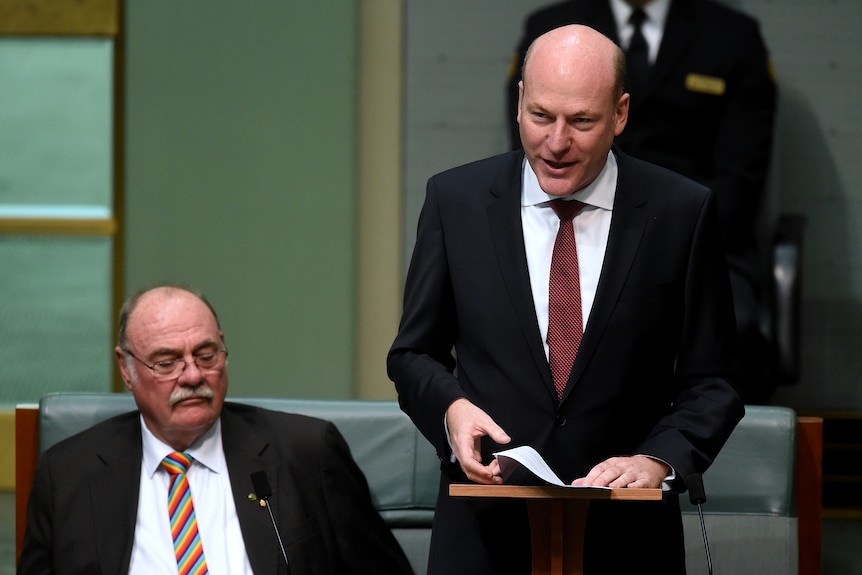 A bald man in a black suit and maroon tie stands up in green House of Representatives to deliver a speech