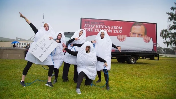 Women dressed in tampon suits stand in front of a billboard with Tony Abbott's face on it