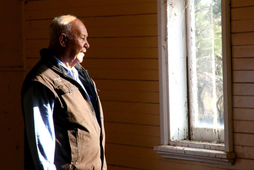 An older man in a brown jacket and blue shirt looks out of the window of a weatherboard house as the sun shines in.