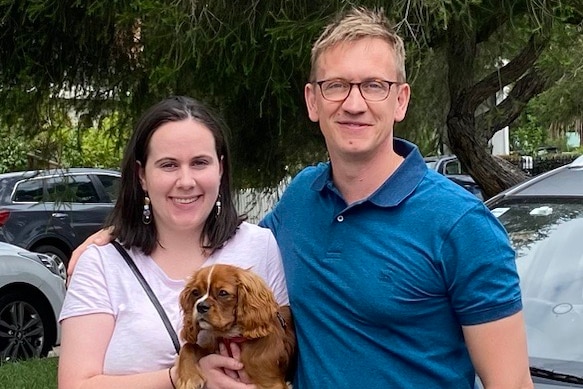 A man poses with his arm around the shoulders of a younger woman who is holding a small dog.