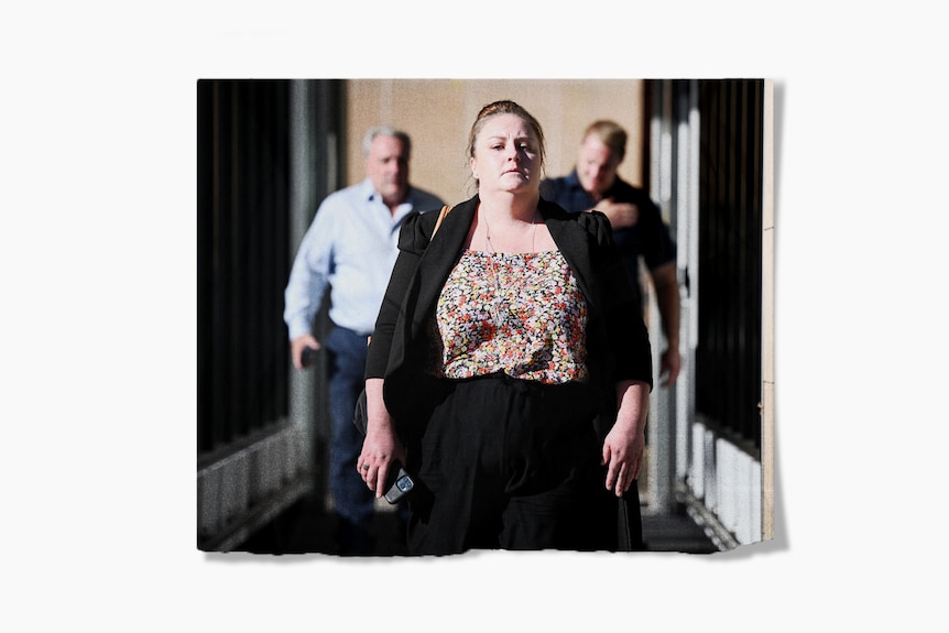 A woman walks outside, towards the camera. She is looking ahead and carrying a phone in her hand.