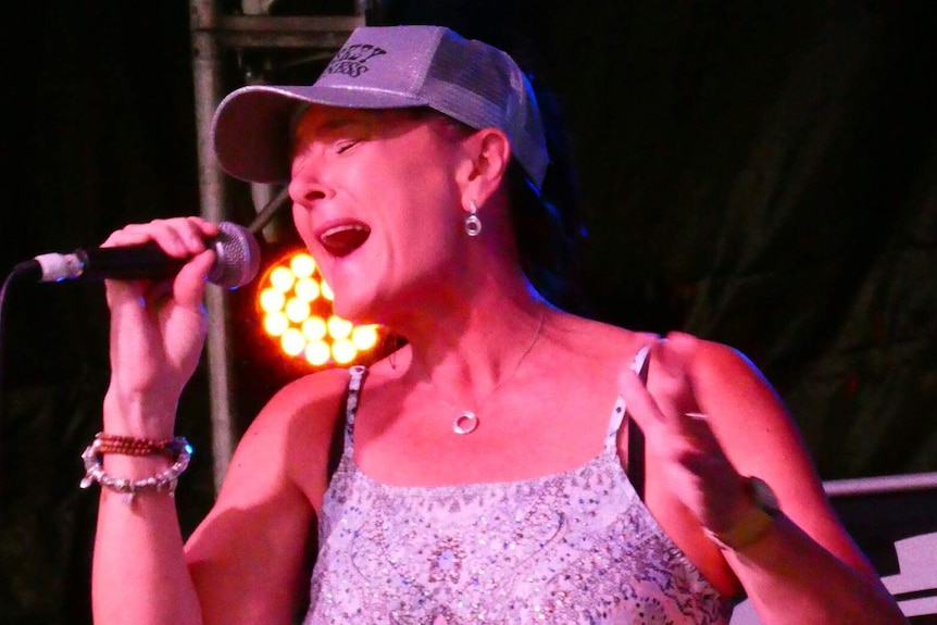 Woman wearing blue jeans, a rhinestone belt and glittering baseball cap sings passionately into a microphone on stage