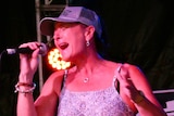 Woman wearing blue jeans, a rhinestone belt and glittering baseball cap sings passionately into a microphone on stage