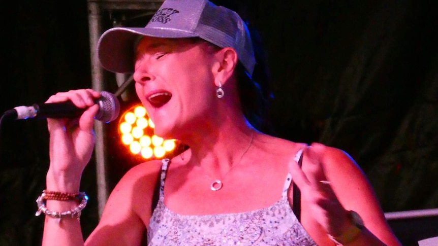 Woman wearing blue jeans, a rhinestone belt and glittering baseball cap sings passionately into a microphone on stage