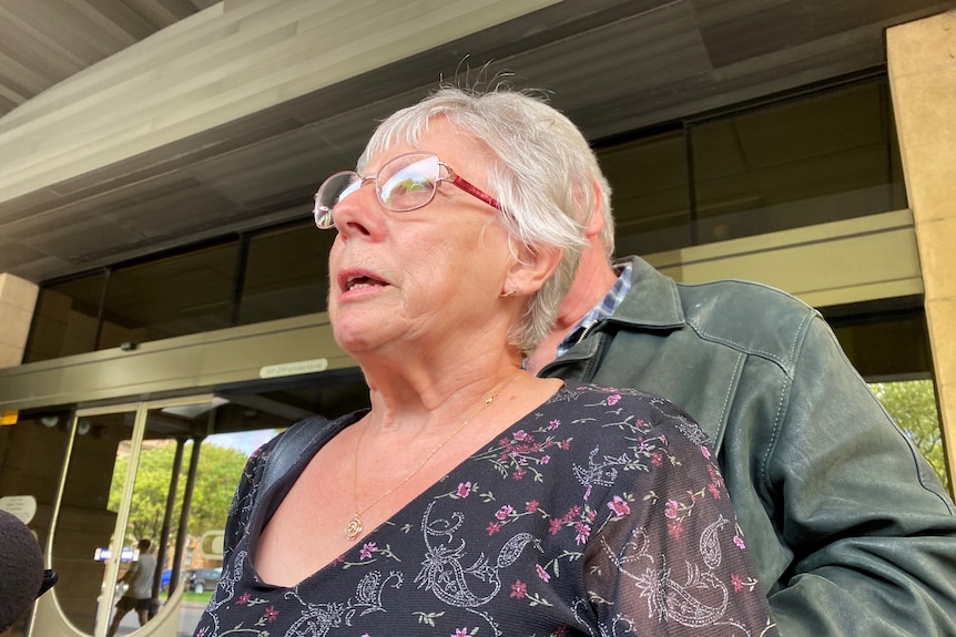 A woman with white hair and red glasses looks off camera