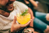 A man smiles while holding an orange mocktail with a sprig of rosemary in it.