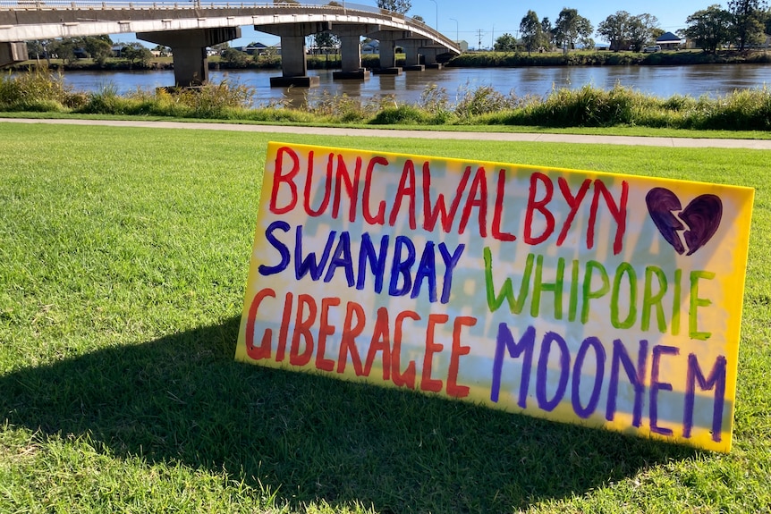 A colourful sign sitting on some grass in front of a bridge over a river.