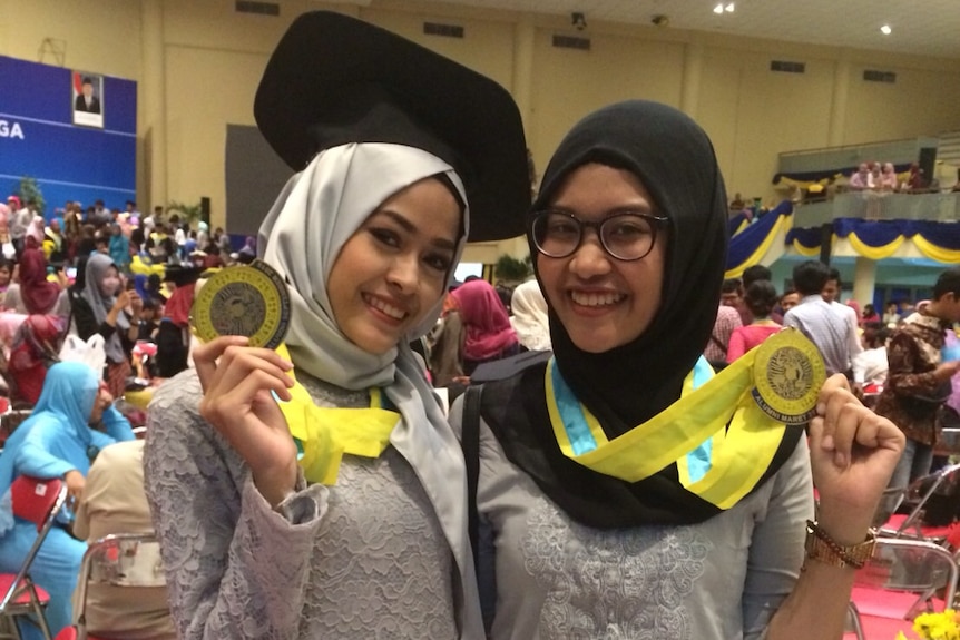 Two women wear formal attire and headscarves while holding medallions at a graduation ceremony