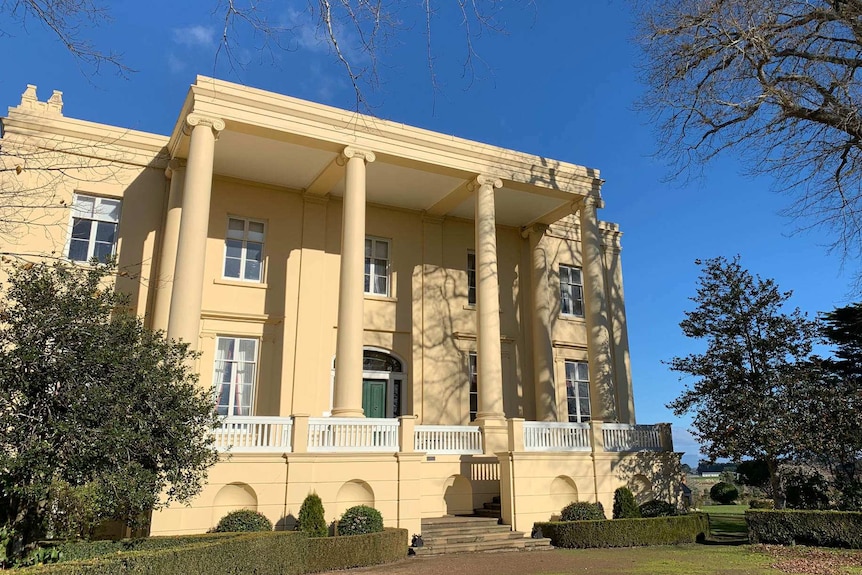 The grand exterior of a two-storey, colonial-era building with large sculpted columns at its entrance