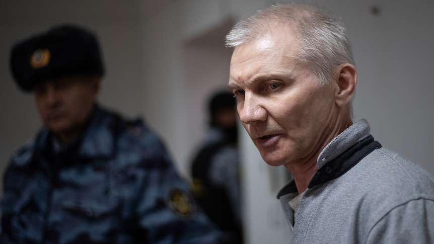 Left is a Russian police officer wearing blue camouflage. Right is an older man with grey hair and a jumper, he looks concerned.
