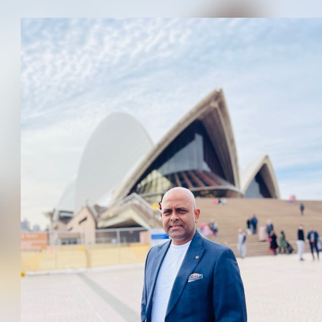 Um homem sorridente e careca, de paletó e camiseta, em frente à Ópera de Sydney. 