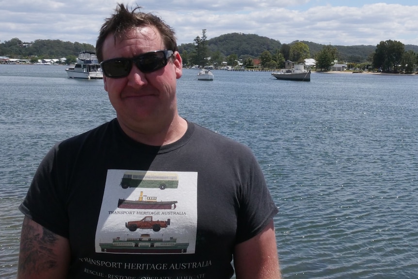 Peter Duffy wears a black tshirt and sunglasses, with the old timber Argos tug moored in the background.