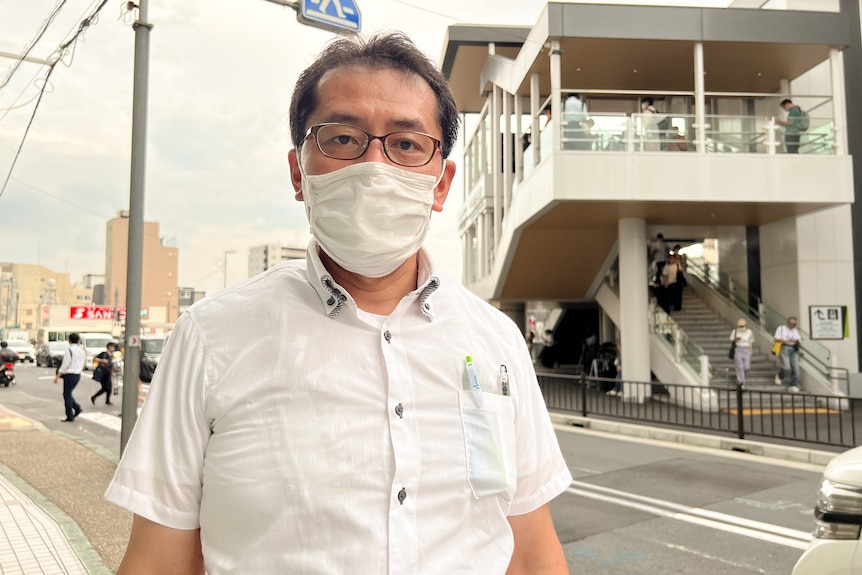 A man wearing a white shirt and white mask stands on a street.
