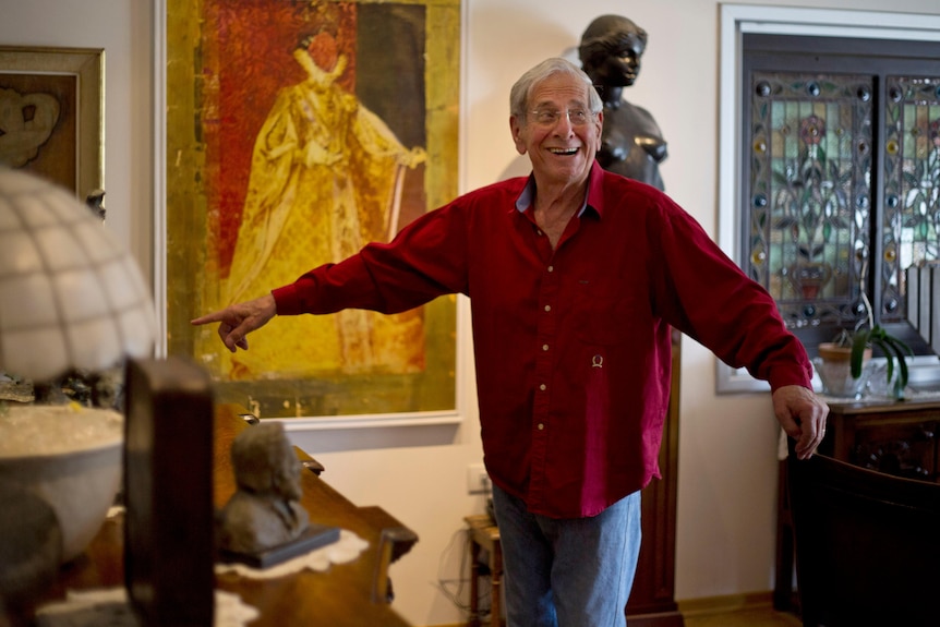 A older man is pictured wearing a red shirt, his arms spread while he smiles. Behind him are some paintings and a statue.