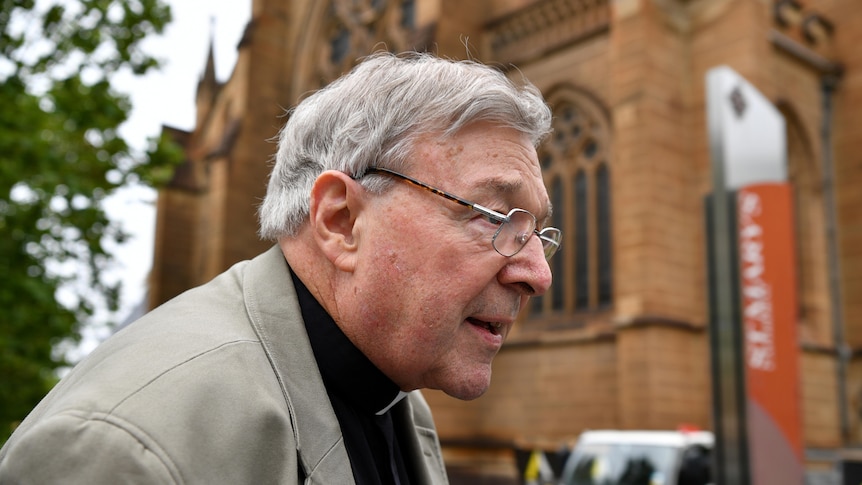 George Pell walks past a sandstone church wearing a jacket