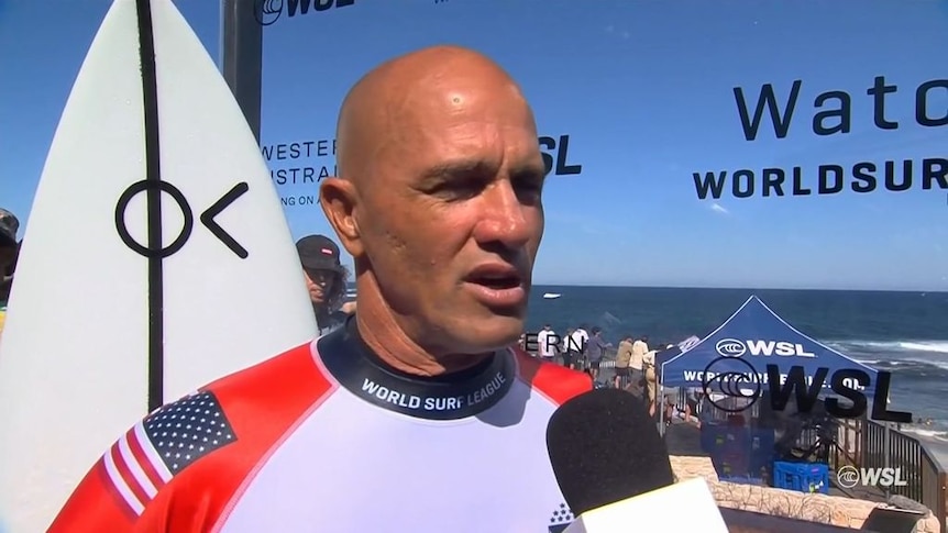 A man with a shaved head gives a television interviewat the beach. There is a surfboard behind him.