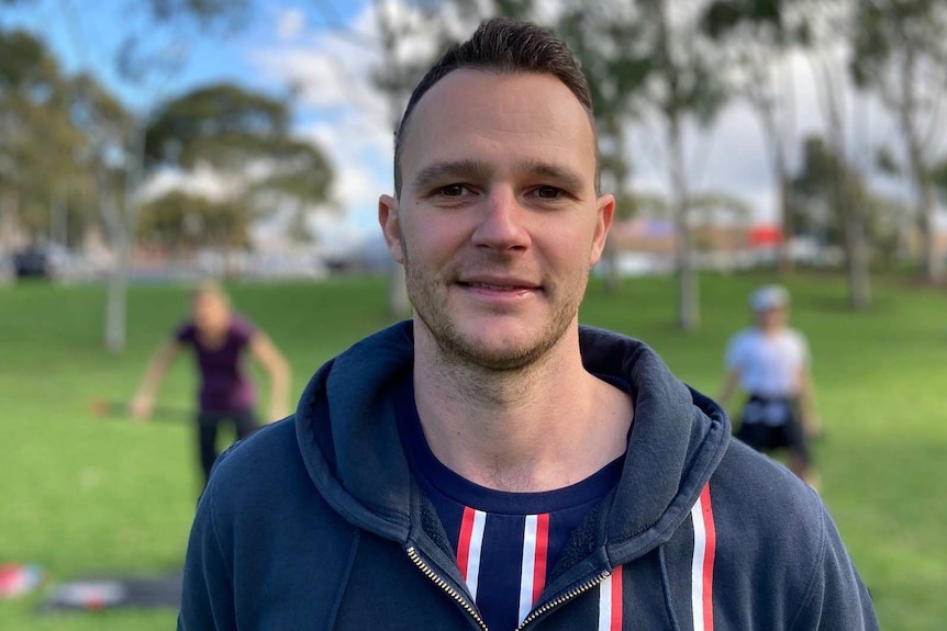 A fitness trainer standing outside in a park with people exercising behind him.