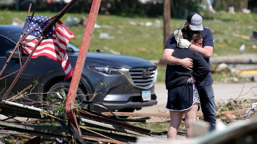 A young man and woman hug beside a wrecked home, on which someone has tied an American flag.
