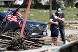 A young man and woman hug beside a wrecked home, on which someone has tied an American flag.